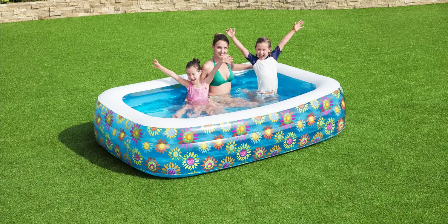 A smiling woman and two young children enjoying a colourful inflatable paddling pool with a bright floral design on green artificial grass in a sunny garden.
